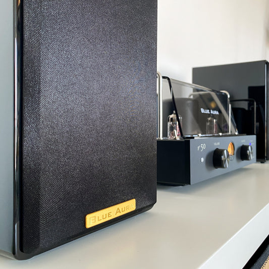Side profile view of the black AT700 left speaker with its fabric grille on and the golden Blue Aura badge.