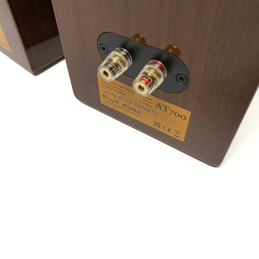 Official close-up angled studio shot of the rear panel of a Blue Aura AT700 Walnut bookshelf speaker, highlighting the premium gold-plated speaker terminals.