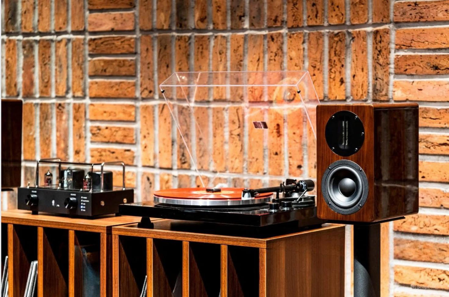 Wide lifestyle view of the Blue Aura Blackline PG2 turntable and a high-gloss Walnut AT700 compact bookshelf speaker without its grille, beautifully arranged on a wooden vinyl record storage cabinet against a rustic brick wall.