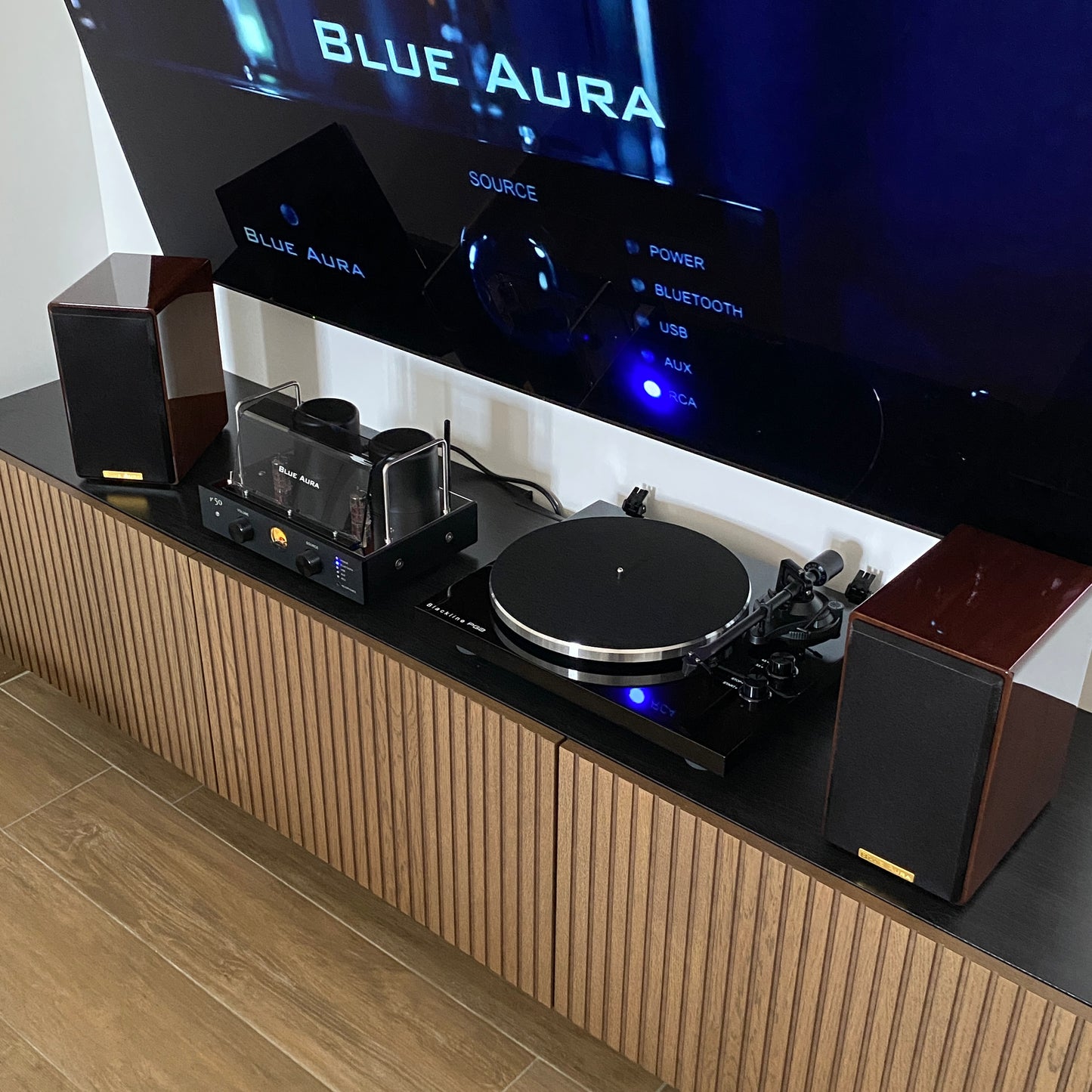 Overview of the Blue Aura Soul Prestige Hi-Fi system with speakers' fabric grilles on and the turned-on amplifier installed underneath the television in a modern living room.