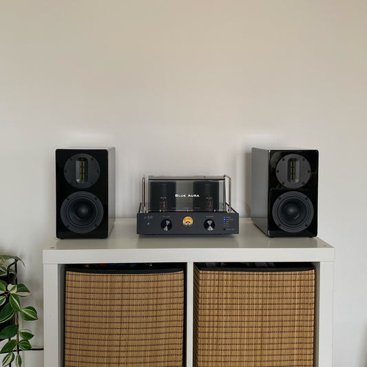 Front view of the Blue Aura v50 hybrid amplifier and black AT700 speakers perfectly centered on a white cabinet with wicker baskets, with the turned-on amplifier visible.