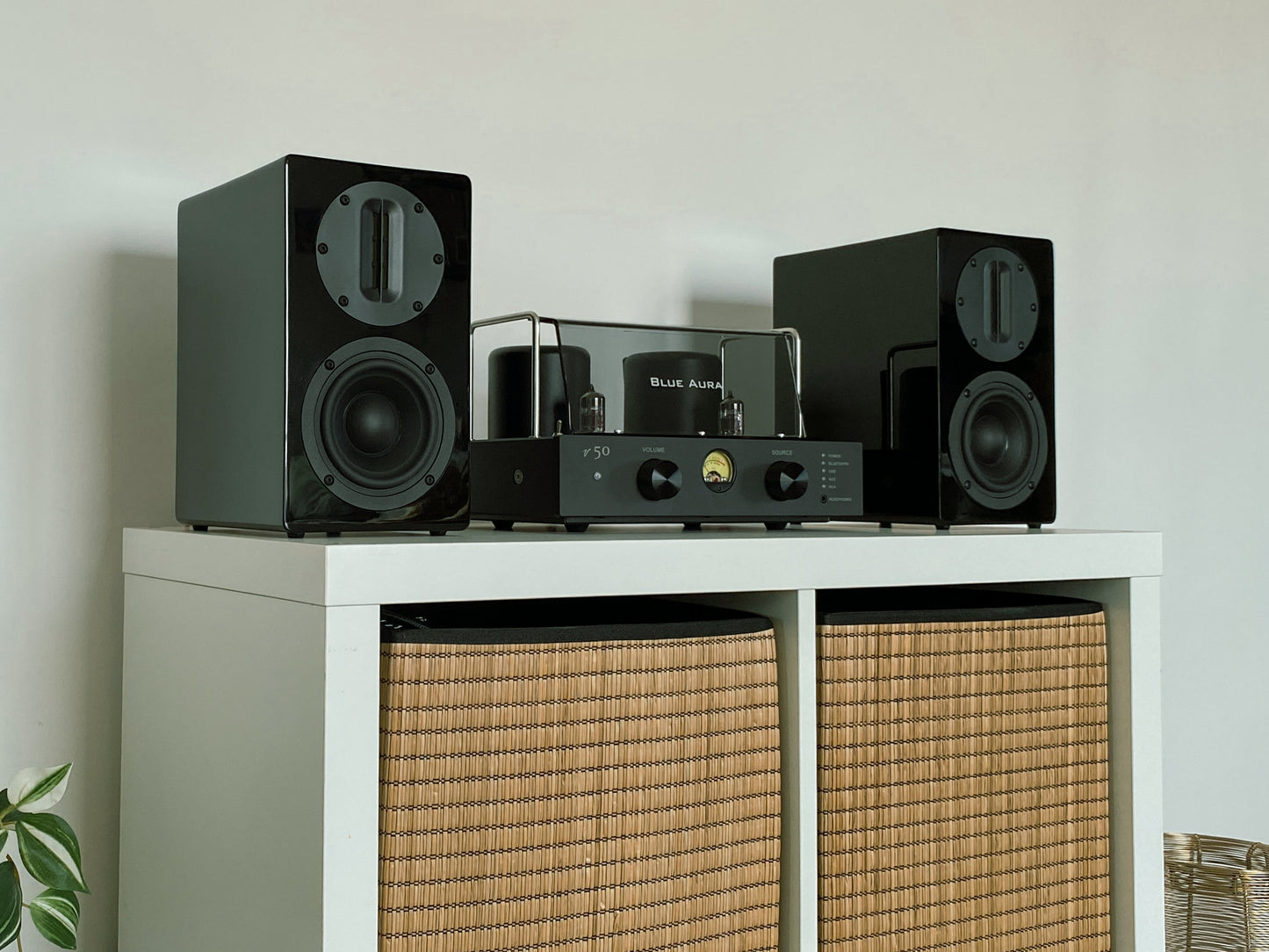 Front lifestyle view of the v50 hybrid amplifier and black AT700 speakers from the Blue Aura Stereo System set up on a white cabinet with wicker baskets.