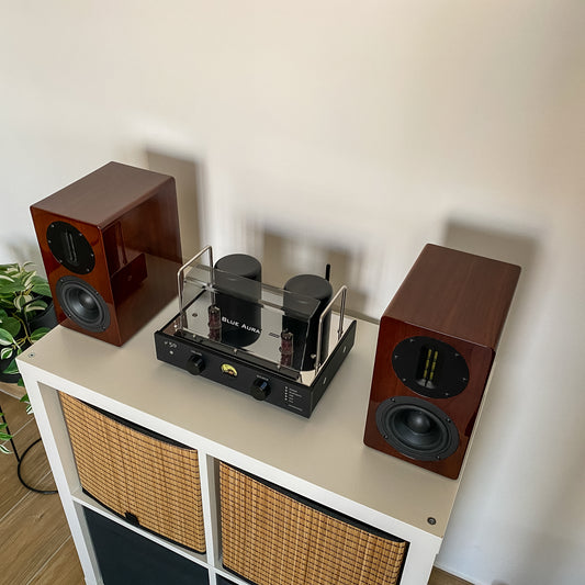 High-angle front lifestyle view of the complete Blue Aura Stereo System on a white cabinet, showcasing the premium high-gloss Walnut AT700 speakers capturing the clear side profile reflection of the v50 hybrid amplifier.