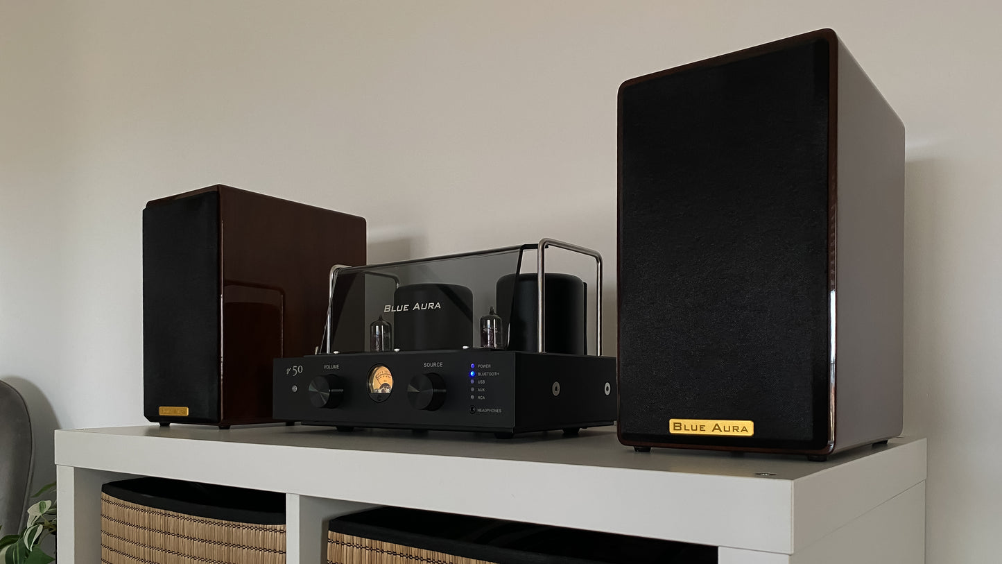 Three-quarter lifestyle view of the high-gloss Walnut AT700 speakers under warm ambient lighting, highlighting the premium gold logo badge on the fabric grilles next to the turned-on v50 amplifier.