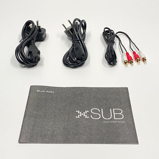 Flat lay studio view of the official setup items included with the Blue Aura X-Sub subwoofer, featuring the UK and EU power cables, RCA cables, and the printed Quick Start Guide booklet.