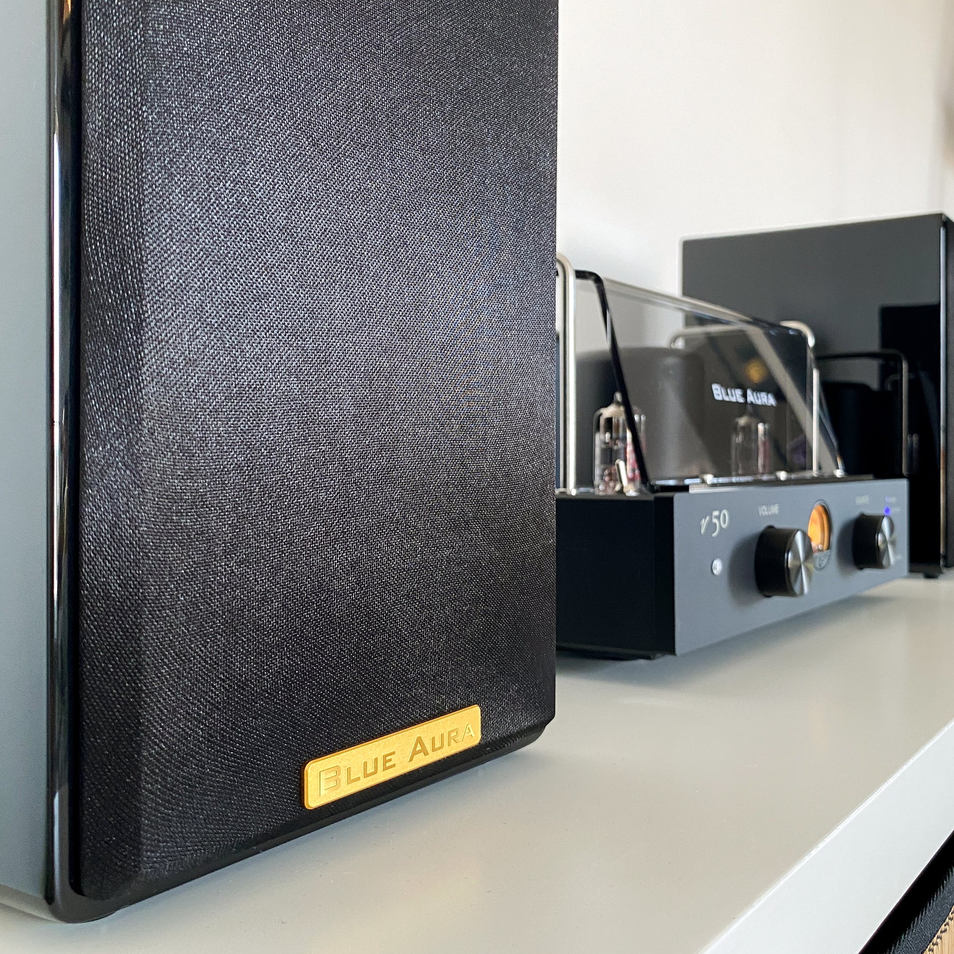 Side profile view of the black AT700 left speaker with its fabric grille on and the golden Blue Aura badge.