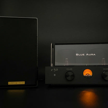 Atmospheric night view of the black AT700 right speaker with its fabric grille on, positioned next to the turned-on v50 amplifier highlighting its illuminated warm-glow VU meter in the dark.