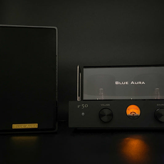 Atmospheric night view of the black AT700 right speaker with its fabric grille on, positioned next to the turned-on v50 amplifier highlighting its illuminated warm-glow VU meter in the dark.