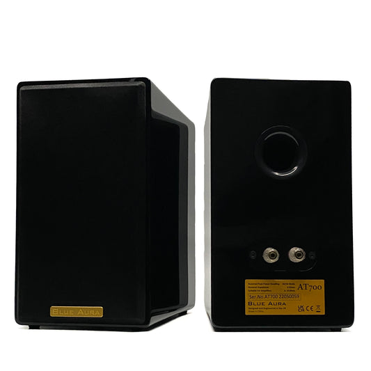 Official studio shot of the Blue Aura AT700 compact bookshelf speakers in high-gloss piano black, displaying one from the front with its grille and the other from the rear showing the circular bass reflex port and gold-plated terminal binding posts.