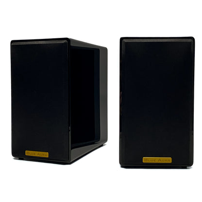 Official studio shot of a pair of Blue Aura AT700 compact bookshelf speakers with their black fabric protective grilles, showcasing the elegant gold metallic brand badges and high-gloss piano black finish.