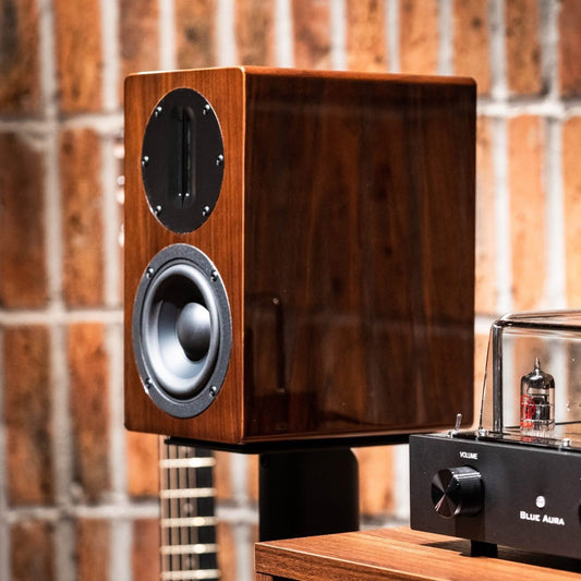 Angled lifestyle view of a single Blue Aura AT700 compact bookshelf speaker, showcasing its premium high-gloss Walnut finish and ribbon tweeter without the fabric grille against a brick wall.