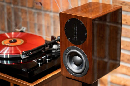 Close-up lifestyle view of a single Blue Aura AT700 compact bookshelf speaker, showcasing its premium high-gloss Walnut finish and ribbon tweeter without the fabric grille against a brick wall.