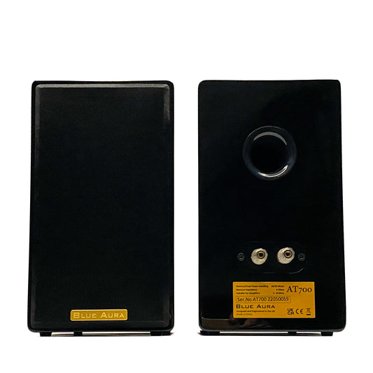Official front and rear studio shot of the Blue Aura AT700 compact bookshelf speakers in high-gloss piano black, displaying one with its grille and the other showing the back panel.