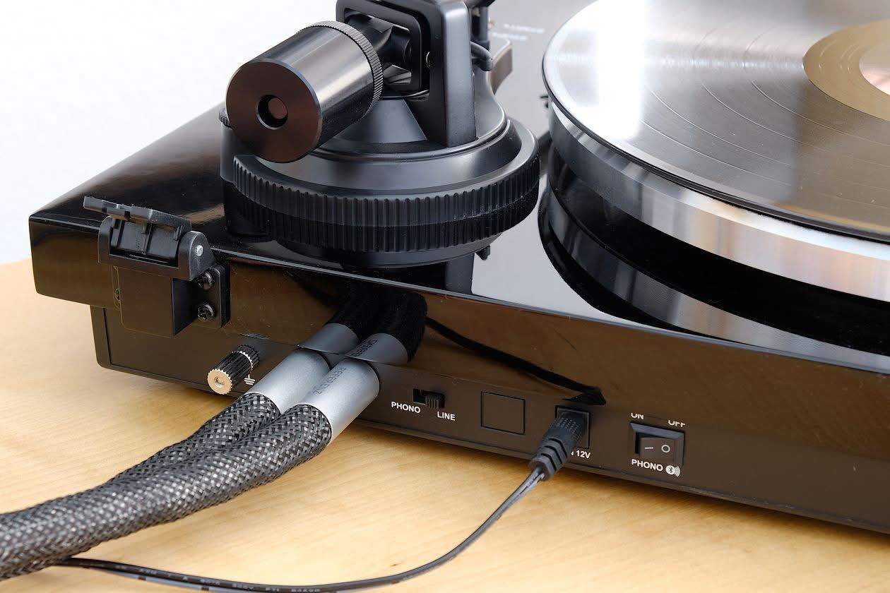 Detailed review close-up from the German HiFi-IFAs test, highlighting the rear panel connections and phono-line preamp switch of the Blackline PG2 turntable, shown with high-end braided RCA cables from the test laboratory.