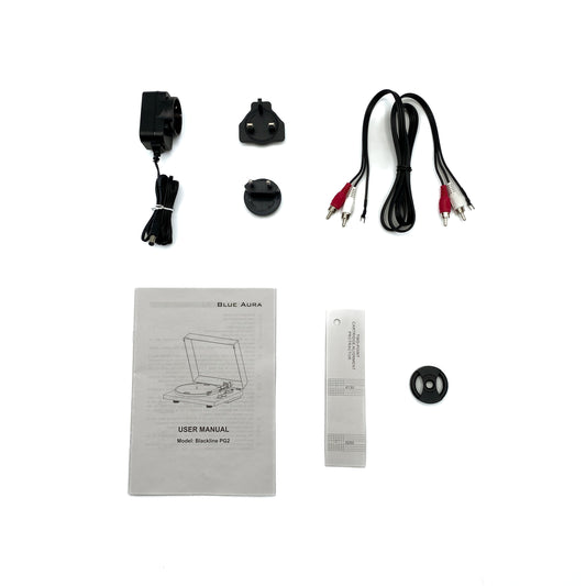 Flat lay studio view of the official setup items included with the Blue Aura Blackline PG2 turntable, featuring the user manual, multi-plug power supply, RCA cables, cartridge alignment protractor, and 45 RPM adapter.