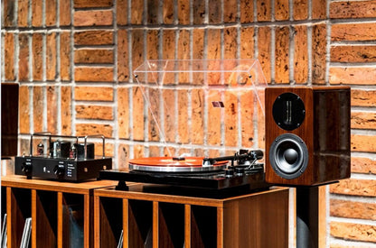 Wide lifestyle view of the Blue Aura Blackline PG2 turntable and a high-gloss Walnut AT700 compact bookshelf speaker without its grille, beautifully arranged on a wooden vinyl record storage cabinet against a rustic brick wall.