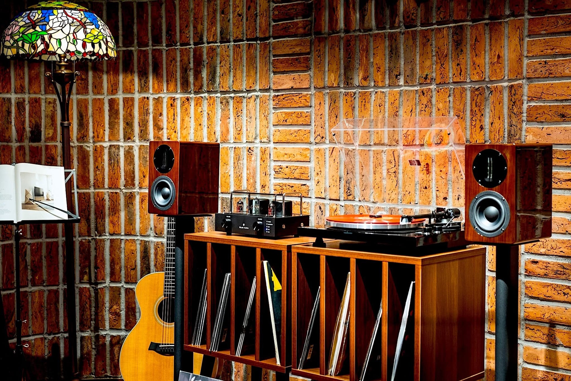 Wide lifestyle view displaying the Blue Aura Blackline PG2 turntable and a pair of high-gloss Walnut AT700 compact bookshelf speakers on universal floor stands, arranged with a wooden storage cabinet against a brick wall.