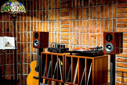 Wide lifestyle view displaying the Blue Aura Blackline PG2 turntable and a pair of high-gloss Walnut AT700 compact bookshelf speakers on universal floor stands, arranged with a wooden storage cabinet against a brick wall.