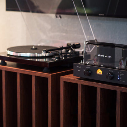 Ambient lifestyle view of the Blue Aura Core System, showcasing the turned-on v50 hybrid amplifier with its prominent red JJ Electronics tube logos and yellow VU meter, next to the Blackline PG2 turntable with its clear dust cover opened on wooden audio furniture.