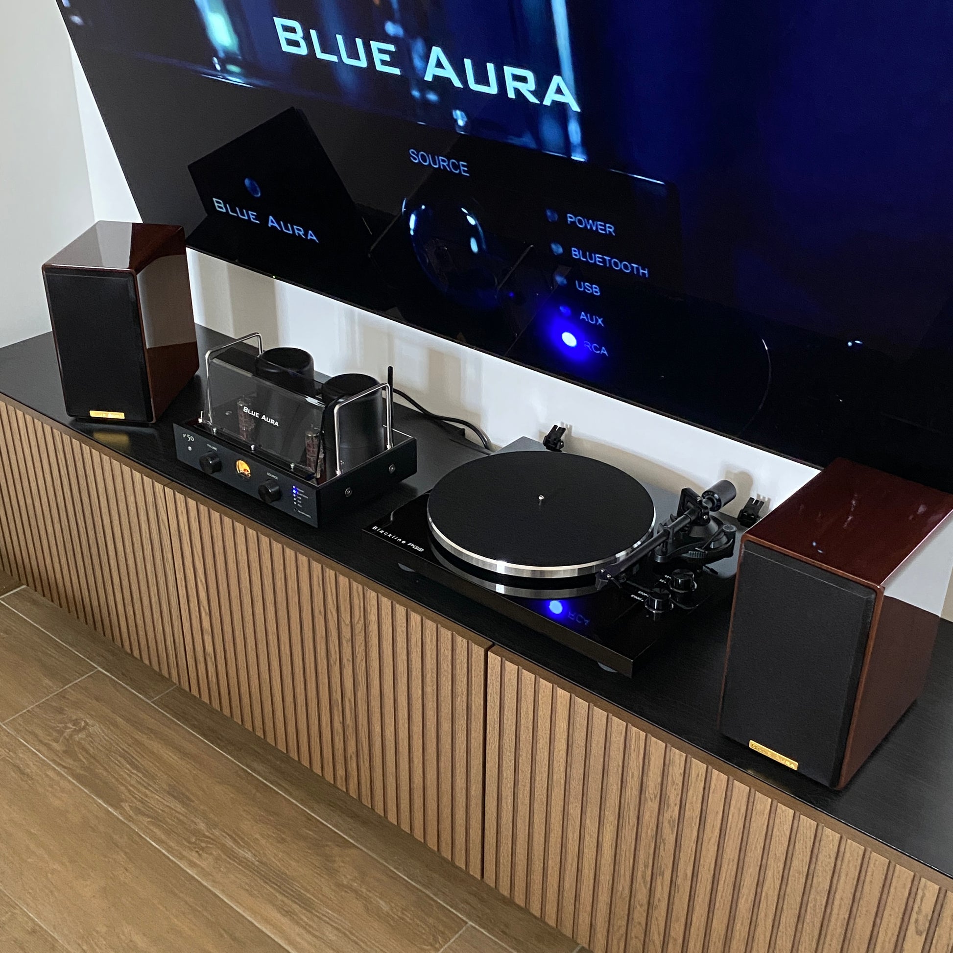 Overview of the Blue Aura Soul Prestige Hi-Fi system with speakers' fabric grilles on and the turned-on amplifier installed underneath the television in a modern living room.