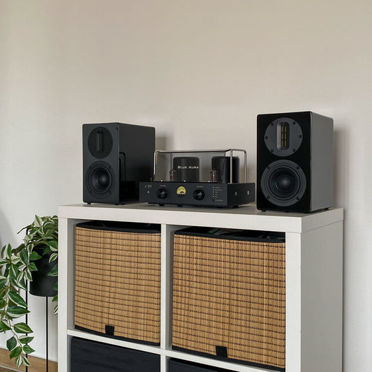 Angled view of the Blue Aura v50 hybrid amplifier and black AT700 speakers set up on a white cabinet with wicker baskets.