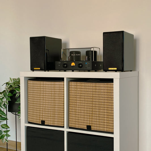 Lifestyle view of the Blue Aura v50 hybrid amplifier and black AT700 speakers set up on a white cabinet with wicker baskets, showing the turned-on amplifier and the speakers with their fabric grilles on.
