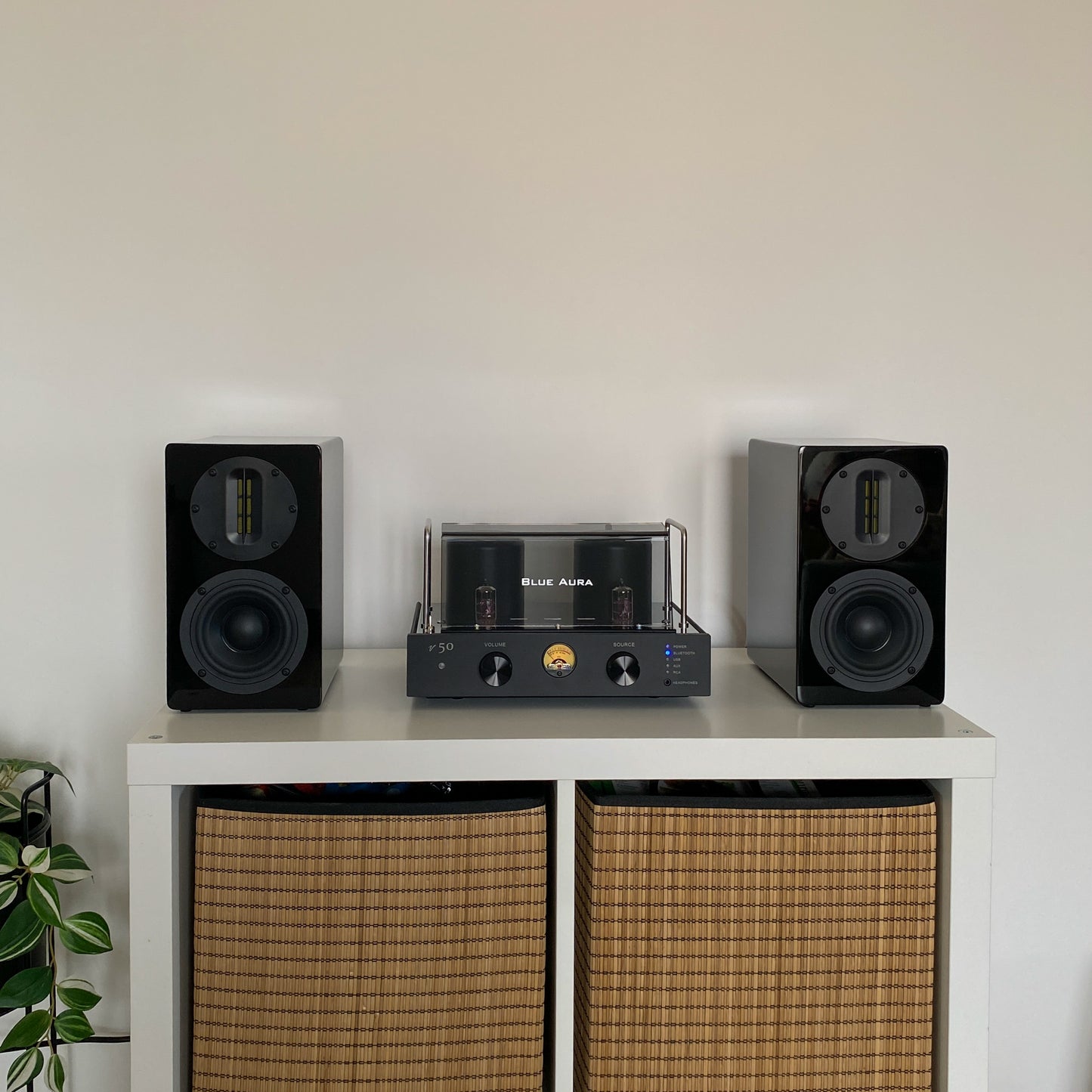 Front view of the Blue Aura v50 hybrid amplifier and black AT700 speakers perfectly centered on a white cabinet with wicker baskets, with the turned-on amplifier visible.