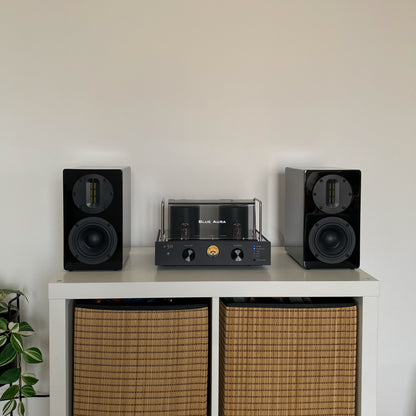 Front view of the Blue Aura v50 hybrid amplifier and black AT700 speakers perfectly centered on a white cabinet with wicker baskets, with the turned-on amplifier visible.