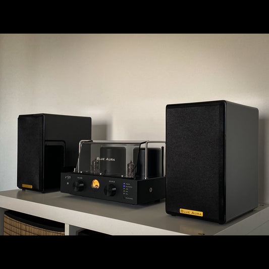 Ambient close-up front view of the black AT700 speakers with their fabric grilles on and the turned-on v50 hybrid amplifier from the Blue Aura Stereo System, arranged on a white cabinet.