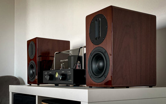 Three-quarter lifestyle view of the v50 hybrid amplifier and high-gloss Walnut AT700 speakers from The Blue Aura Stereo System on a white cabinet.