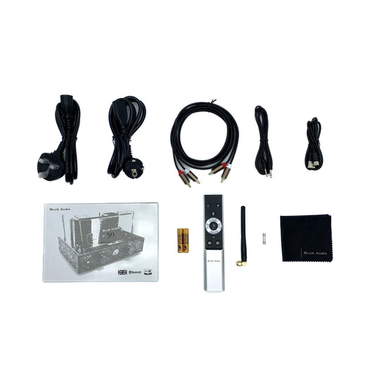 Flat lay studio view of the official setup items included with the Blue Aura v50 hybrid amplifier, featuring the user manual, brushed aluminium remote control, power cables, RCA cords, Bluetooth antenna, and microfibre cleaning cloth.