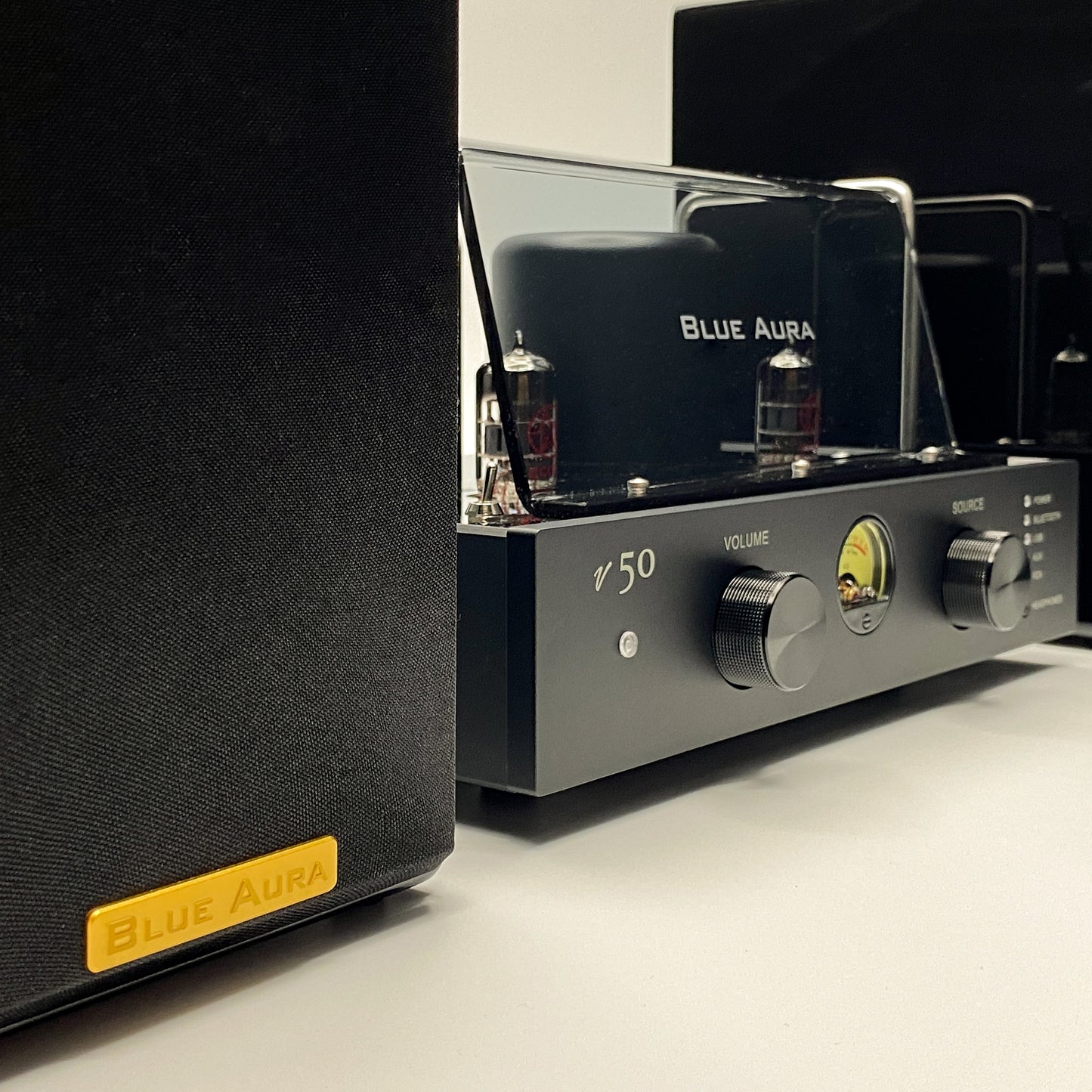 Angled front view of the Blue Aura v50 hybrid valve amplifier positioned next to the black AT700 speaker with its fabric grille and golden badge in the foreground.