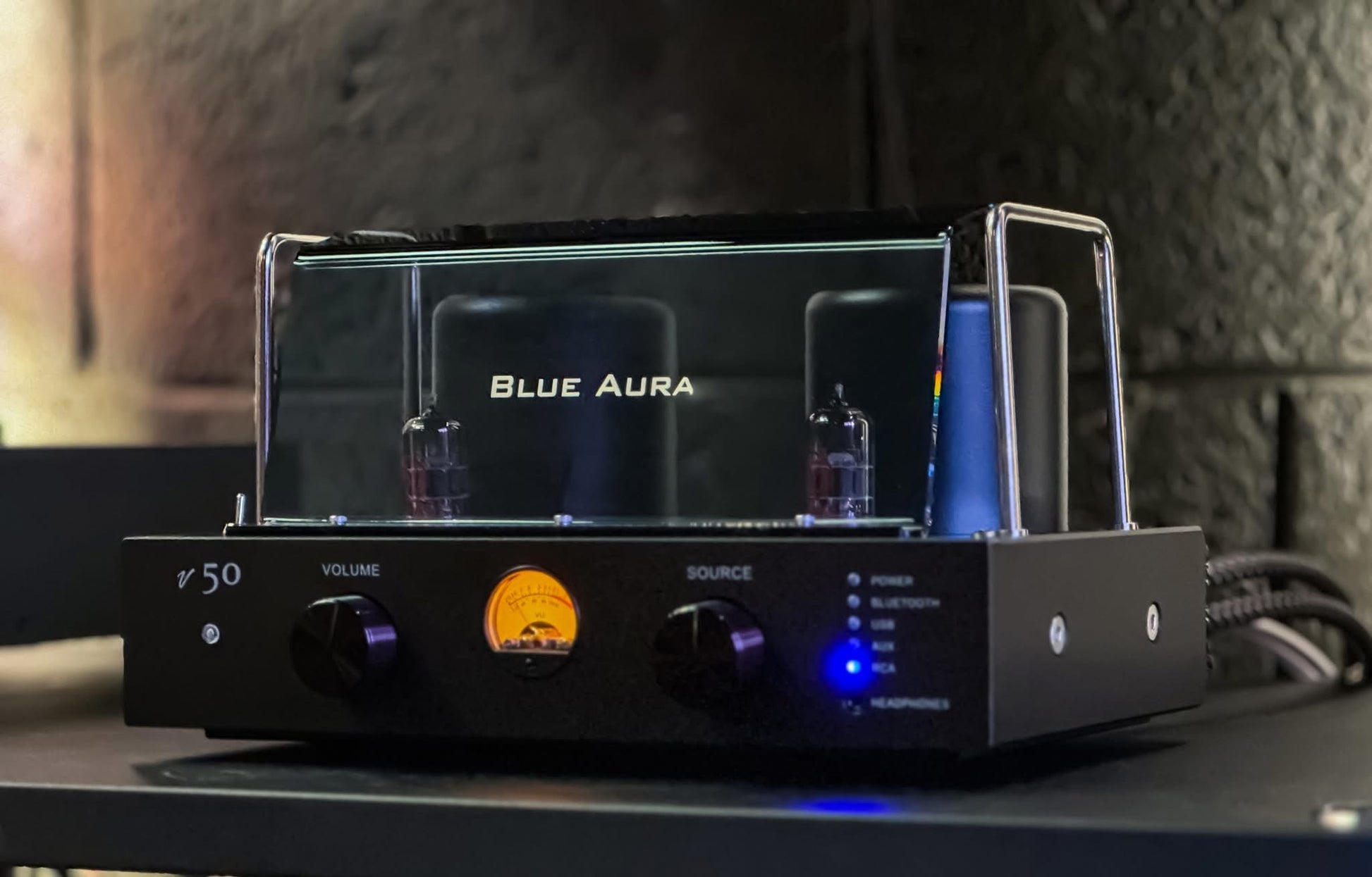 Three-quarter lifestyle view of the turned-on Blue Aura v50 hybrid valve amplifier under warm ambient lighting, showcasing its illuminated VU meter, glowing blue LED indicator, and branded protective plexiglas.