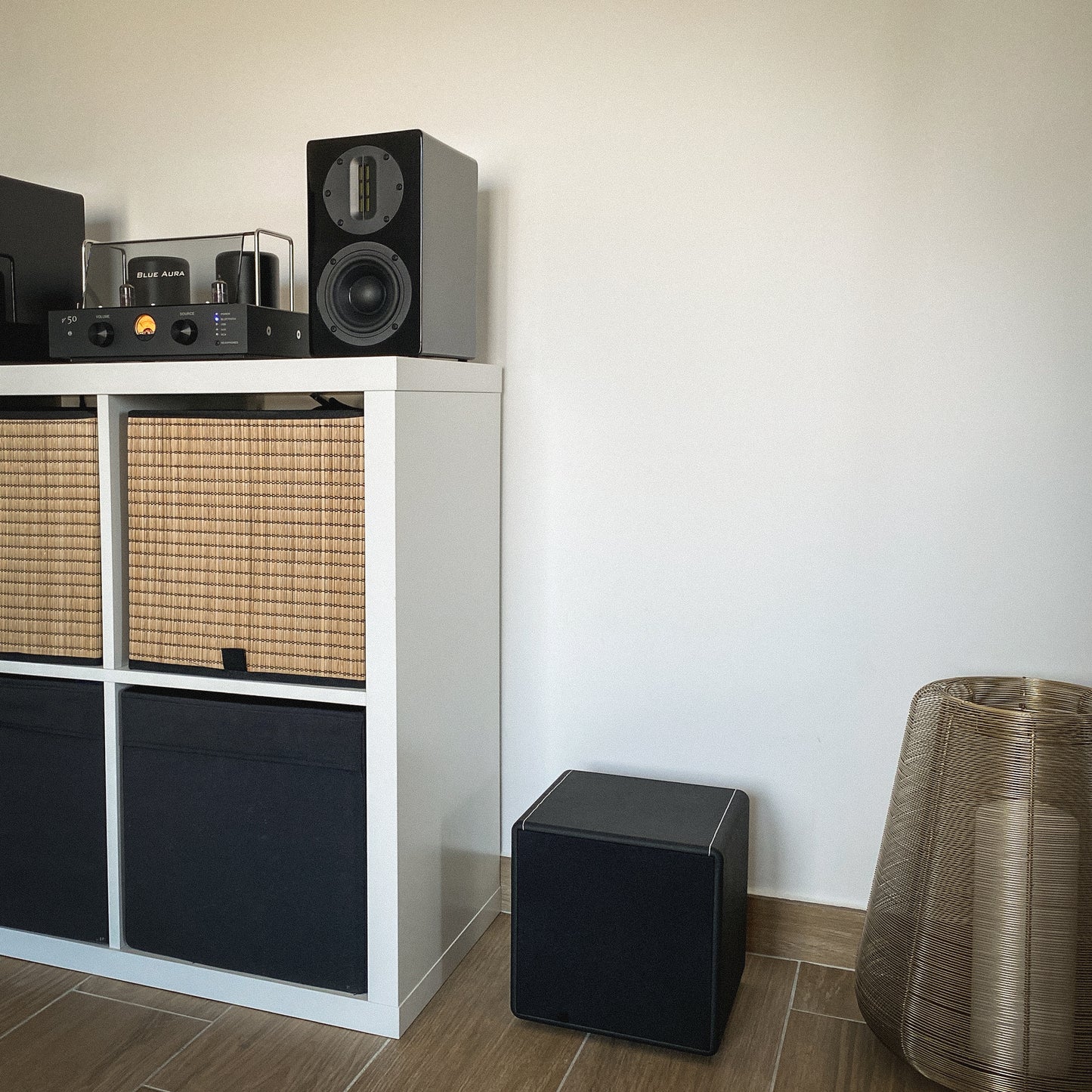 Close-up of the Blue Aura X-Sub subwoofer on the floor with fabric grille, with the v50 stereo system and black AT700 speakers in the background.