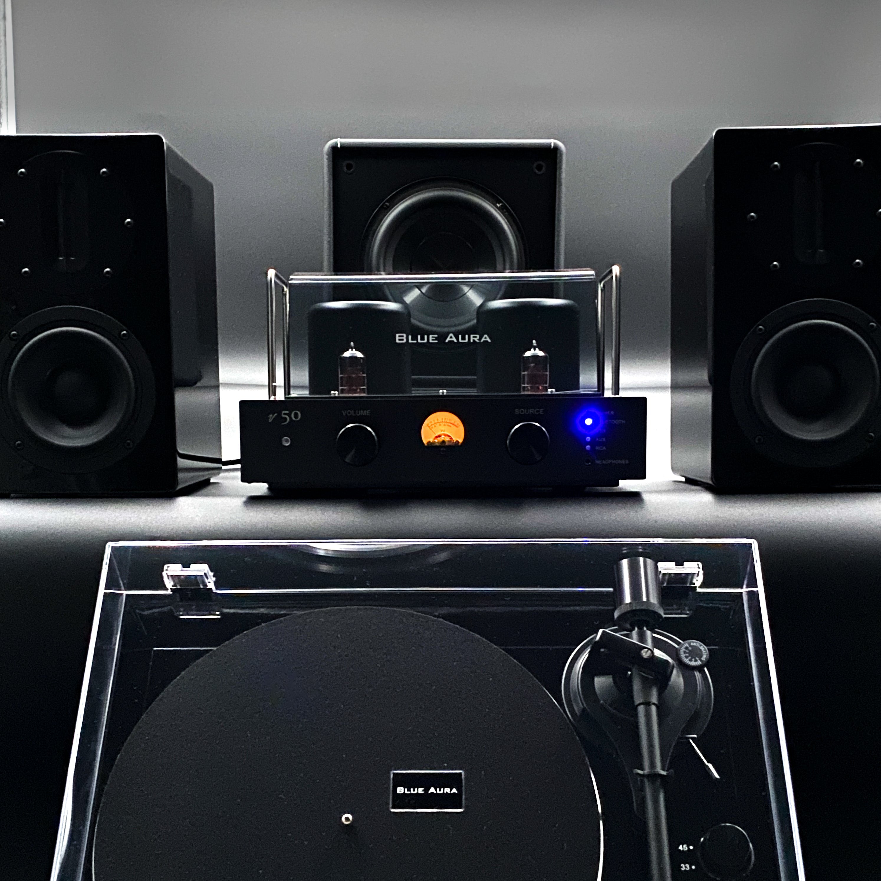 Official studio view of the complete Blue Aura Blackline stereo system pack on a moody grey background, showcasing the turned-on v50 amplifier, AT700 black speakers, X-Sub subwoofer, and Blackline PG2 turntable.
